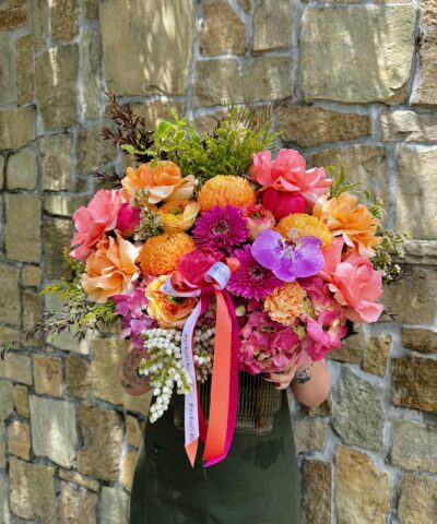 Tropical Crush is a vibrant floral arrangement in a glass vase featuring hot pink peonies, tangerine roses, fuchsia orchids, and golden chrysanthemums, finished with The Lush Lily signature ribbons.