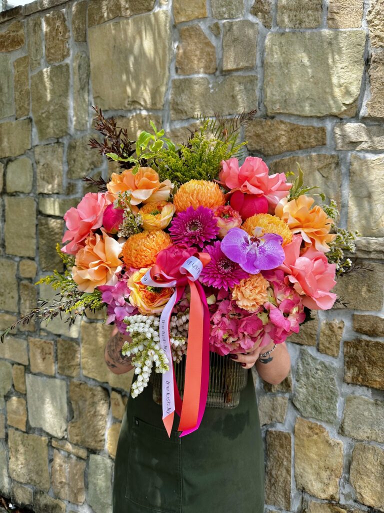 Tropical Crush is a vibrant floral arrangement in a glass vase featuring hot pink peonies, tangerine roses, fuchsia orchids, and golden chrysanthemums, finished with The Lush Lily signature ribbons.