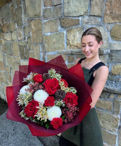 Luxurious Christmas bouquet featuring red roses, white disbuds, red carnations, pinecones and festive foliage, wrapped in deep burgundy paper — handcrafted by The Lush Lily florist in Brisbane.