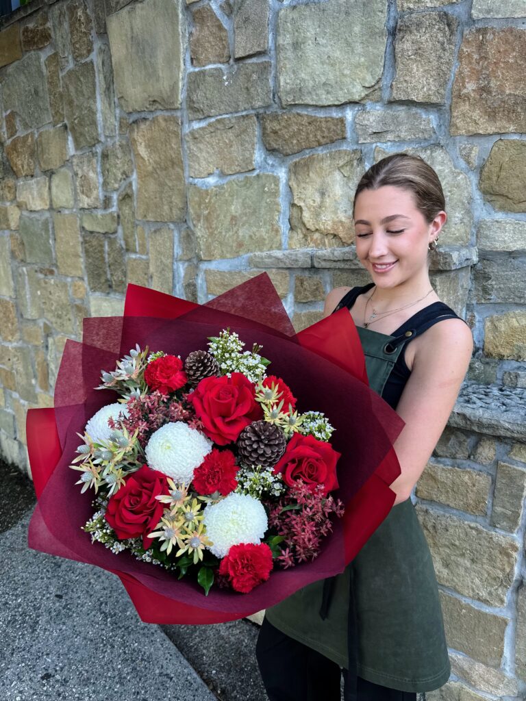 Luxurious Christmas bouquet featuring red roses, white disbuds, red carnations, pinecones and festive foliage, wrapped in deep burgundy paper — handcrafted by The Lush Lily florist in Brisbane.