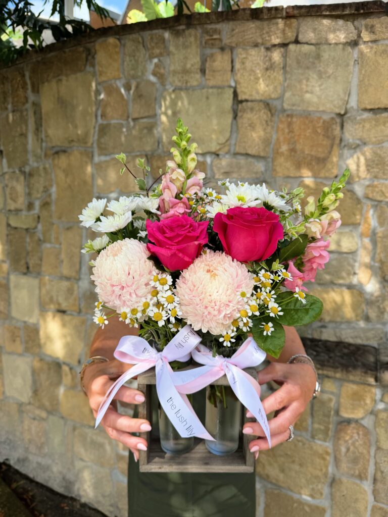 Meet our Blush Garden Duo — a sweet and stylish floral arrangement designed to capture the beauty of a soft, romantic garden in full bloom. Thoughtfully arranged in two elegant glass vases, this charming duo is presented in a rustic wooden holder and finished with The Lush Lily’s signature satin ribbon for that extra luxe touch. Featuring a delicate mix of blush chrysanthemums, premium pink roses, dainty chamomile-style daisies, and seasonal filler flowers, the Blush Garden Duo is all about soft textures, feminine tones, and effortless beauty. Each bloom is carefully selected fresh from market to ensure a high-quality, long-lasting arrangement that feels both whimsical and refined. This unique double-vase design makes it incredibly versatile — perfect as a centrepiece, bedside styling, or a thoughtful gift that feels a little different from your traditional bouquet. The two vases can be styled together for a fuller look or separated to spread the love across multiple spaces. Whether you’re celebrating a special moment or simply sending a little love, this arrangement is designed to suit a wide range of occasions: 💗 Perfect for: * Mother’s Day * Birthdays * Get Well Soon gestures * Thank You gifts * New baby celebrations * Thinking of You moments * Just because At The Lush Lily, we pride ourselves on creating floral designs that feel as though they’ve been freshly picked from a garden. While we always aim to replicate the colours and style pictured, flowers may vary slightly due to seasonal availability and market freshness. Rest assured, any substitutions will always be made with blooms of equal or greater value, maintaining the overall look, feel, and colour palette of this design. Each Blush Garden Duo is handcrafted with care by our talented florists and beautifully wrapped, making it ready to gift and guaranteed to impress. Enjoy same-day flower delivery across Brisbane when you order before our cut-off time — because beautiful flowers should never have to wait.