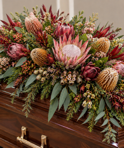 Forever in Nature Australian native casket spray with pink king proteas, golden banksias, burgundy foliage and cascading eucalyptus on a coffin.